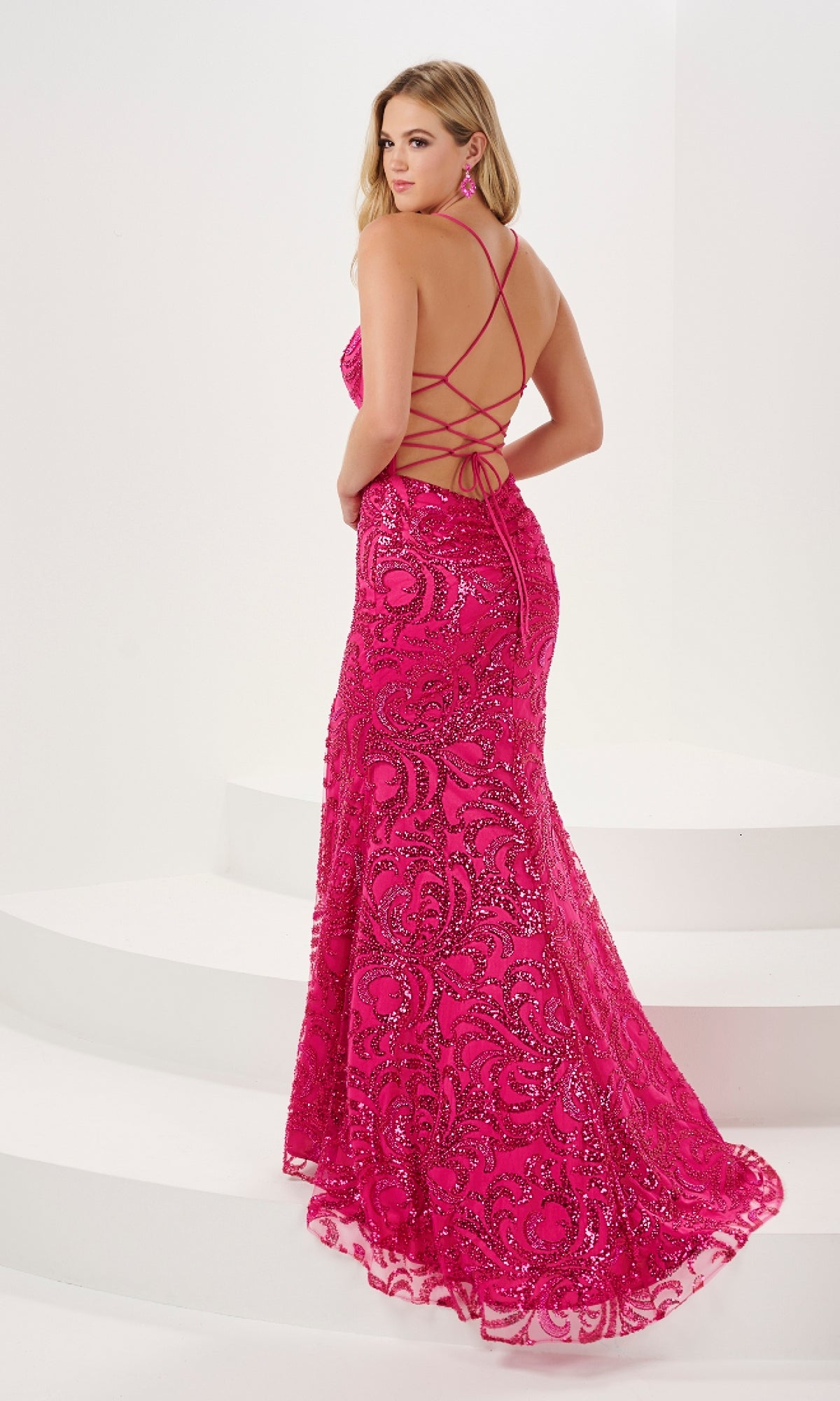 A woman stands on white steps wearing the Long Prom Dress 14186 by Panoply, a bright pink sequin gown with a lace-up back, strappy open details, and a fitted silhouette.